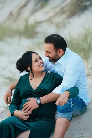 An expectant couple sit together on a sandy dune and smile at each other on an Adelaide beach.
