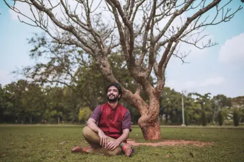 A man in a red polo sits cross-legged and smiles on the grass beneath a bare tree in an Adelaide park.