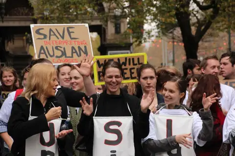 Women in sign-lettered aprons smile and sign in Auslan at a Deaf community march in Melbourne.