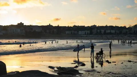 At sunset on Bondi Beach a wheelchair user and surfers stand at the water's edge with the Bondi skyline behind.