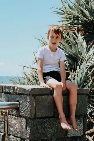 A cheerful boy in a white t-shirt sits on a stone wall in front of pandanus palms at Burleigh Heads.