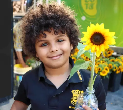 A curly-haired child in a school polo grins at the camera while holding a sunflower in a bottle at a Brisbane market.