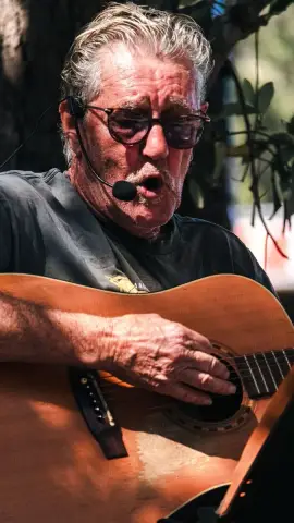 A grey-haired street performer in sunglasses sings into a headset mic and plays an acoustic guitar outdoors.
