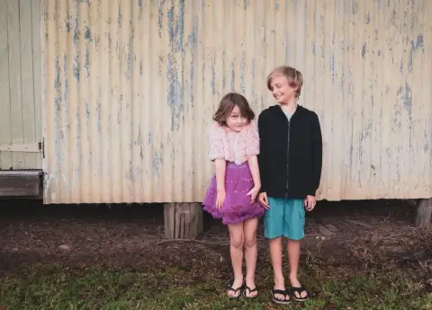 A girl in a purple tutu and a boy in a black jacket stand side by side in front of a weathered shed in Currumbin.