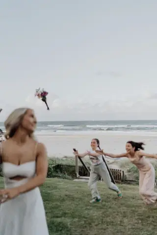 Guests in light dresses run and laugh as the bridal bouquet flies through the air at a beach wedding on the Gold Coast.