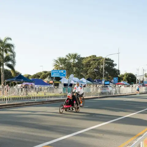 A handcyclist and a cyclist ride along a palm-lined road past event tents at a community sports event on the Gold Coast.