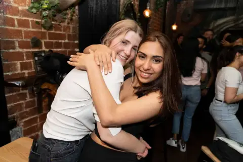 Two friends hug and grin at the camera in a bar on a night out in Melbourne.