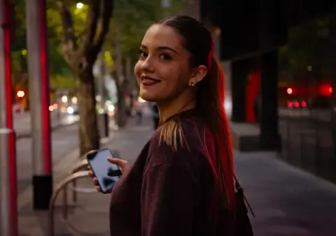 A young woman holding a phone looks back over her shoulder and smiles on a Melbourne street in the evening.