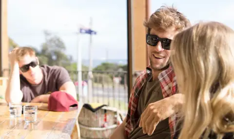 Three friends in sunglasses laugh together at an outdoor cafe table with an Australian flag flying behind them.
