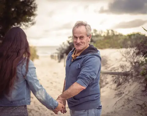 An older man in a hoodie smiles at the camera as he holds hands with a woman on a sandy beach path in Perth.