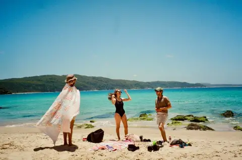 Three friends hang out on a sunny beach at Seal Rocks with turquoise water and a bush headland behind them.