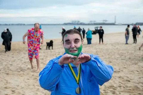 A Special Olympics athlete with a medal and green face paint makes a heart shape with his hands on the beach at the Polar Plunge in South Melbourne.