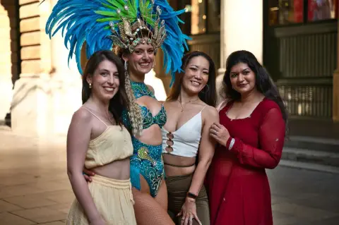 Four women, including a dancer in a blue feathered costume, smile together at an outdoor gathering in Sydney.