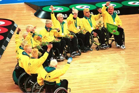 The Australian wheelchair rugby team in yellow jackets cheer with raised arms and medals on court at the 2000 Sydney Paralympic Games.