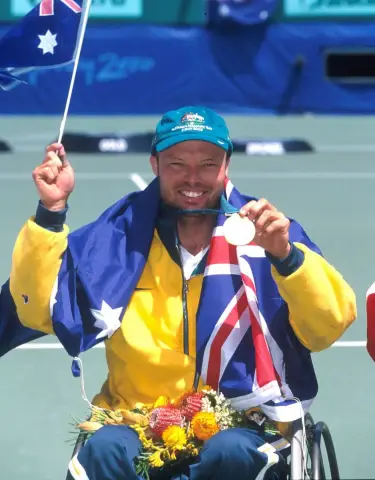Wheelchair tennis player David Hall smiles on the podium draped in an Australian flag, holding flowers and a flag, at the 2000 Sydney Paralympic Games.